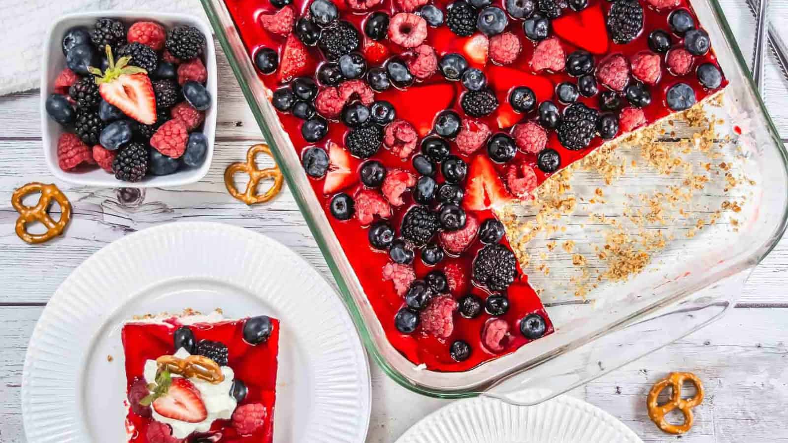 A slice of berry-topped dessert with a pretzel crust, whipped cream, and a strawberry half sits on a white plate. A glass dish with more dessert and a bowl of mixed berries are in the background.