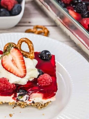 A slice of berry-topped dessert with a pretzel crust, whipped cream, and a strawberry half sits on a white plate. A glass dish with more dessert and a bowl of mixed berries are in the background.