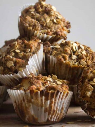 A stack of six oat-topped muffins in white paper liners sits on a wooden surface with a neutral background.