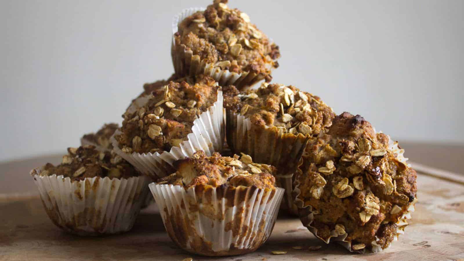 A stack of six oat-topped muffins in white paper liners sits on a wooden surface with a neutral background.