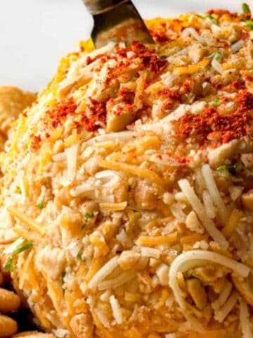 A round cheese ball coated with shredded cheese, chopped nuts, and spices sits on a platter, surrounded by a ring of round crackers. A knife is inserted into the top of the cheese ball.