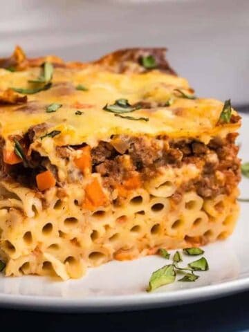 A square slice of baked pasta casserole with ground meat, carrots, and melted cheese, garnished with chopped herbs, sits on a white plate next to a fork.