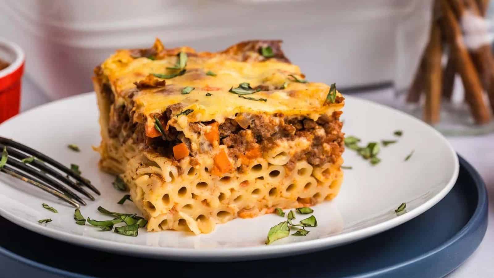 A slice of pasta casserole with ground meat and melted cheese on a white plate, garnished with chopped herbs.