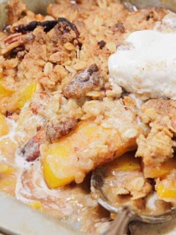 A baking dish filled with peach crisp topped with a scoop of melting vanilla ice cream. The crisp has a crumbly topping and chunks of peaches, with a serving spoon resting inside the dish.