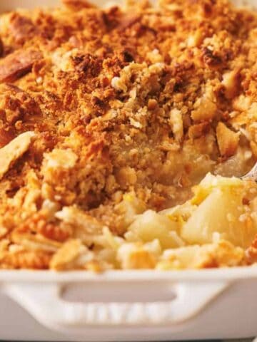 A rectangular white baking dish filled with golden brown apple crumble, with a serving spoon inside and a portion taken out. A spoon and napkin are placed nearby on the table.