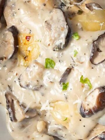 A close-up of ravioli pasta topped with a creamy mushroom sauce, garnished with chopped herbs and grated cheese, served in a white bowl.