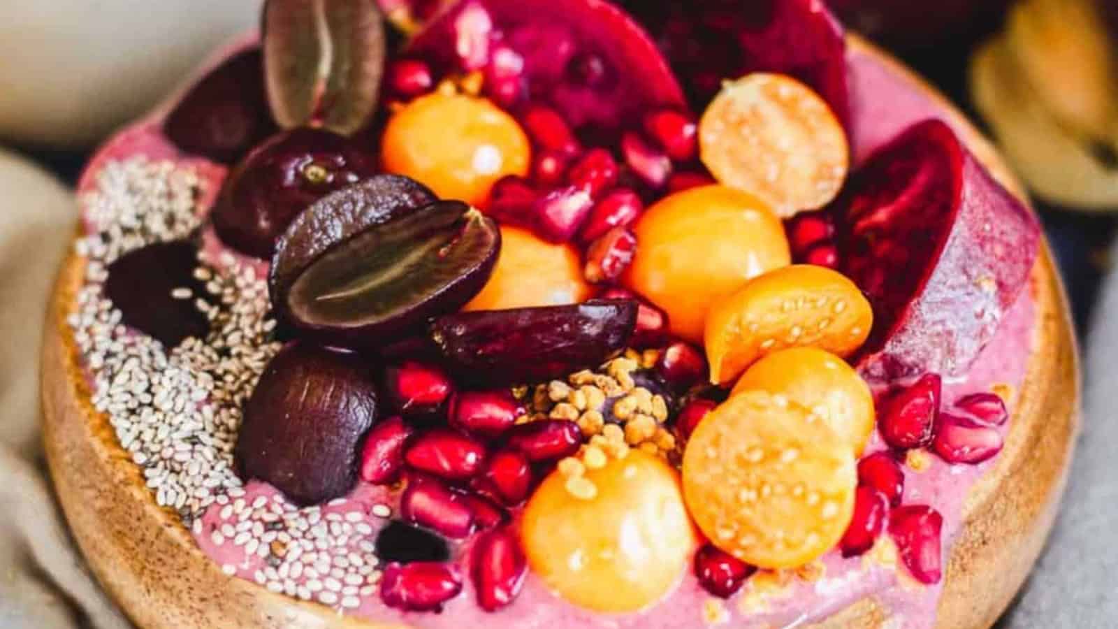A smoothie bowl topped with halved grapes, sliced plums, pomegranate seeds, golden berries, chia seeds, and bee pollen, placed next to a vintage spoon on a beige cloth.