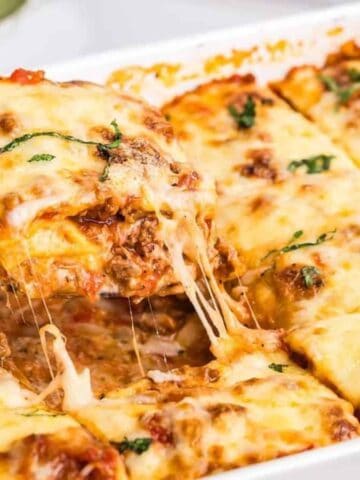 A close-up of a baked lasagna in a white dish, with a serving being lifted out. The lasagna has layers of pasta, meat sauce, melted cheese, and is garnished with small pieces of fresh herbs.