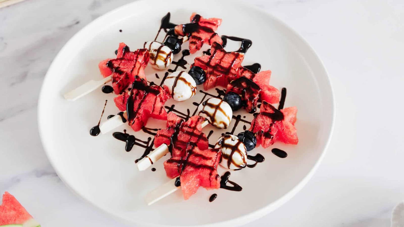 Watermelon and blueberry skewers with mozzarella balls are arranged on a white plate, drizzled with balsamic glaze. The skewers are placed on a marble surface.
