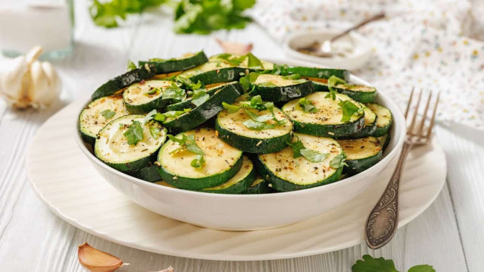 A white bowl filled with sliced Saut&eacute;ed Zucchini with Garlic and Herbs, garnished with chopped herbs, is placed on a white plate with a fork nearby.