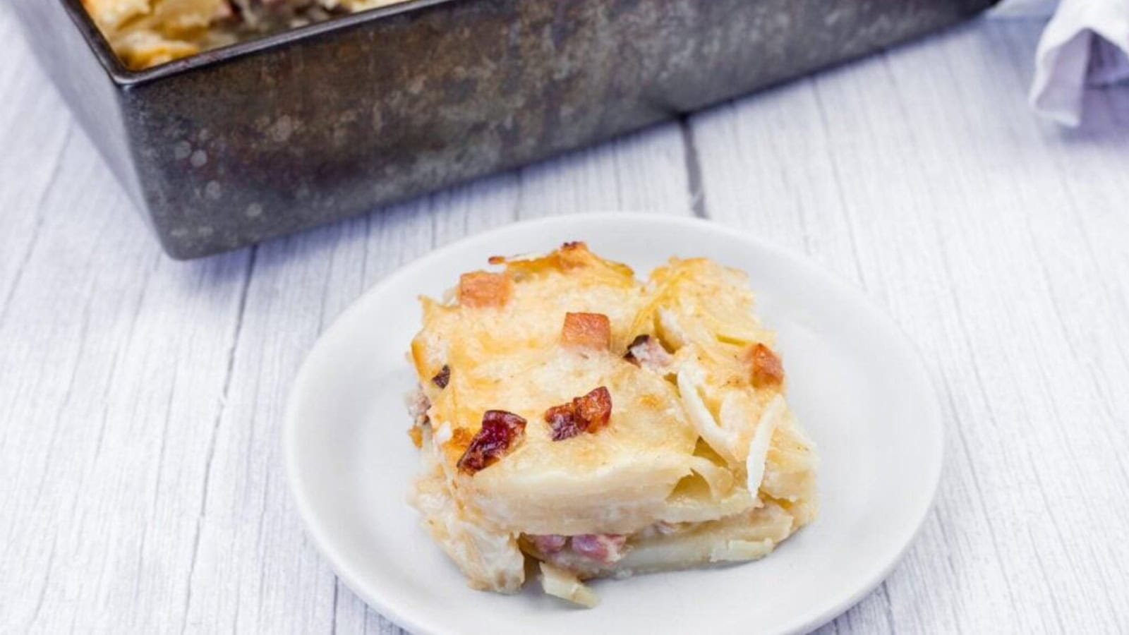 A slice of Scalloped Potatoes with Ham and Cheese is served on a white plate, with the rest of the dish visible in a metal baking pan on a light wooden surface.