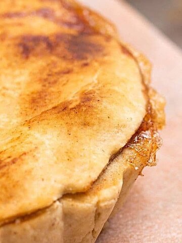 A close-up view of a golden-brown baked pie with a slightly flaky crust and small slits on the top, sitting on a light-colored surface. Only part of the pie is visible in the frame.