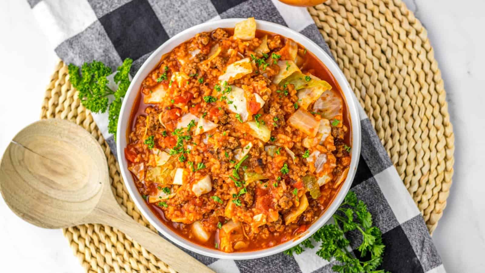 A bowl of stuffed cabbage roll soup with ground meat, cabbage, and tomato sauce, garnished with chopped parsley, sits on a woven placemat next to a wooden spoon.