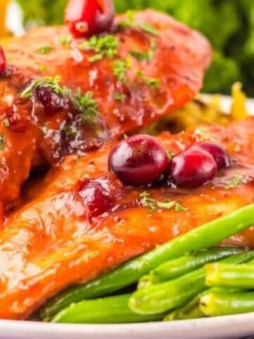 A plate with two pieces of glazed chicken garnished with cranberries, served with green beans and a yellow side dish, possibly cornbread stuffing, on a white plate.