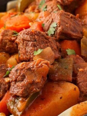 A close-up of a bowl filled with beef stew, featuring chunks of beef, carrots, potatoes, green bell peppers, and fresh herbs, served over a bed of wide egg noodles.
