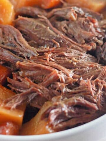 A close-up of a white bowl filled with shredded pot roast, cooked carrots, and chunks of potatoes. The vegetables and meat appear tender and seasoned.