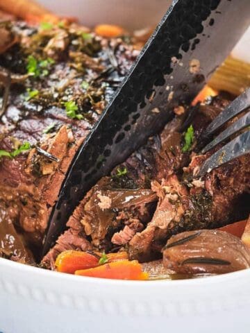 A close-up of a cooked pot roast in a white baking dish with carrots, onions, and celery. A hand uses a fork and knife to cut into the meat. The dish sits on a blue checkered cloth.