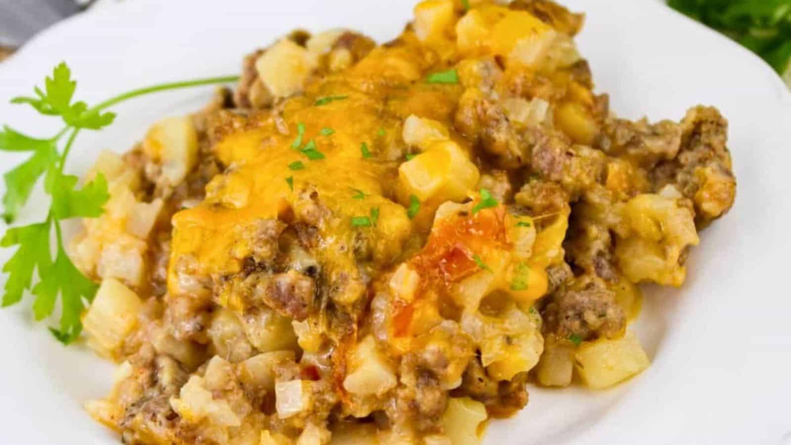 A white plate with a casserole dish, including ground meat, potatoes, and melted cheese, garnished with a parsley sprig.