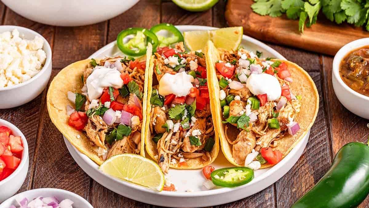 Three chicken tacos with toppings including sour cream, diced tomatoes, onions, jalape&ntilde;os, and cilantro on a white plate, garnished with lime wedges.