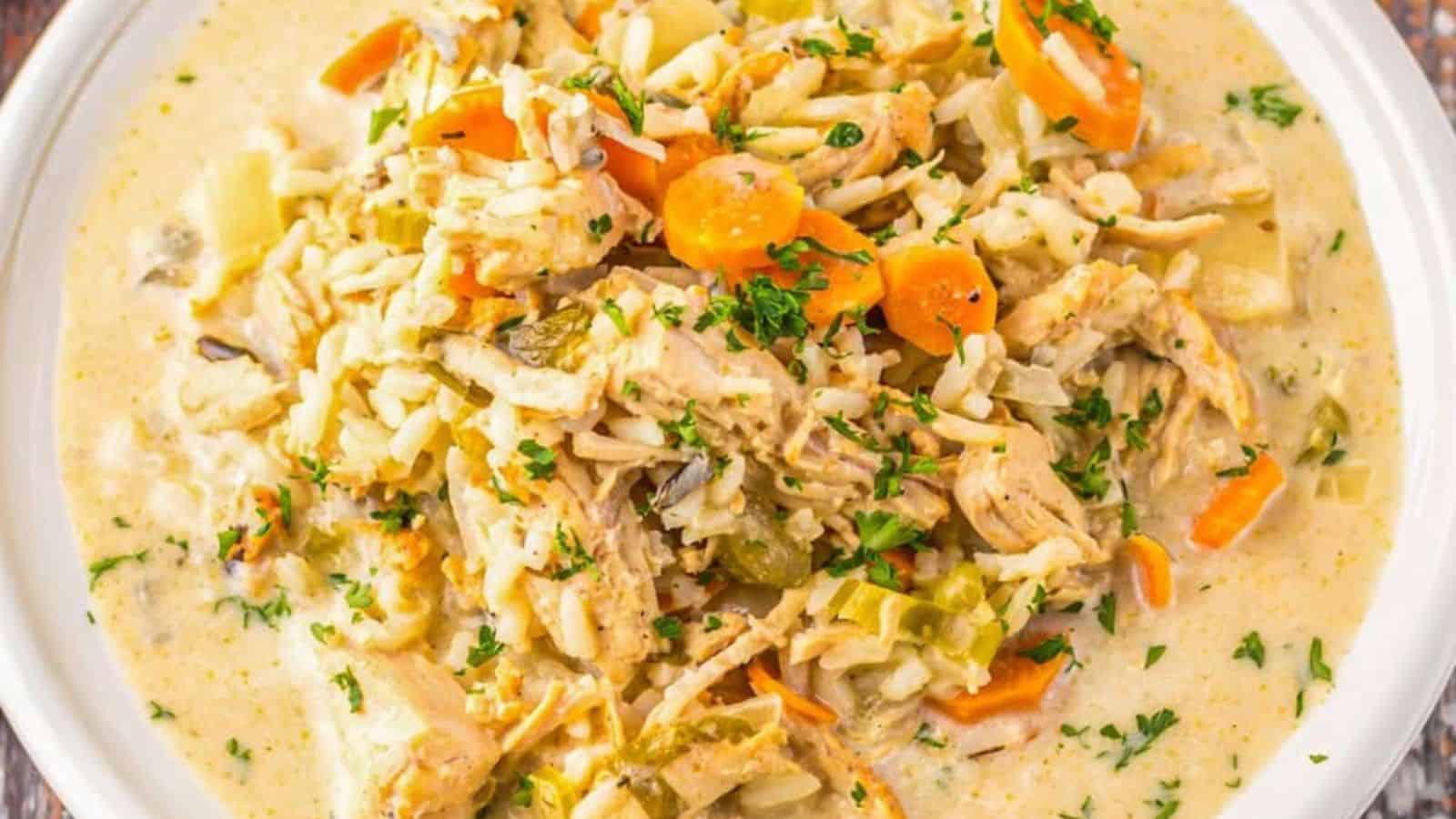 A bowl of creamy chicken and rice soup with shredded chicken, sliced carrots, celery, and herbs, served on a white plate.