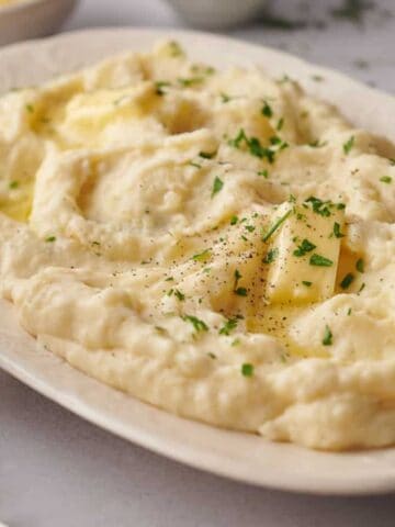 A white oval dish filled with creamy mashed potatoes, topped with melted butter, black pepper, and chopped parsley. A small pitcher and a bowl are in the background on a light surface.