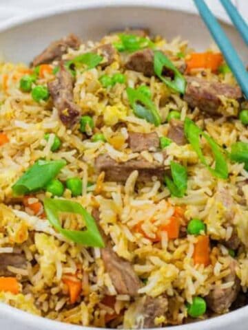 A white bowl filled with fried rice, beef strips, green peas, carrots, scrambled eggs, and green onions. A pair of blue chopsticks rests on the side of the bowl.