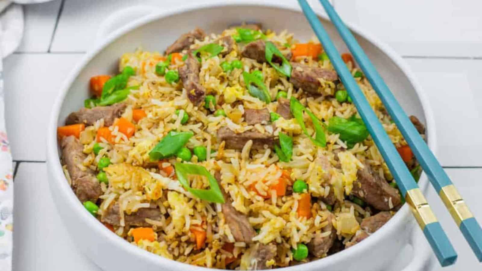 A bowl of steak fried rice served with blue chopsticks on a white tiled surface.