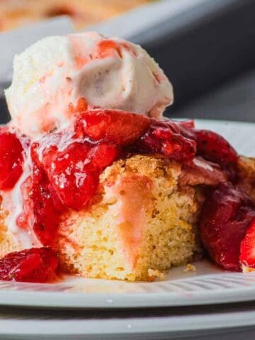 A slice of cake topped with strawberry sauce and a scoop of vanilla ice cream sits on a white plate, with a halved fresh strawberry beside it. A baking tray and whole strawberries are in the background.