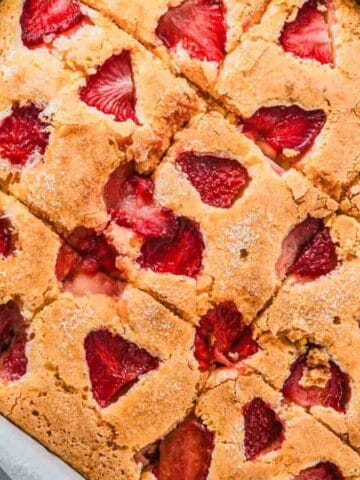 A rectangular baking dish lined with parchment paper holds a golden-brown sheet cake topped with sliced strawberries, cut into square pieces.