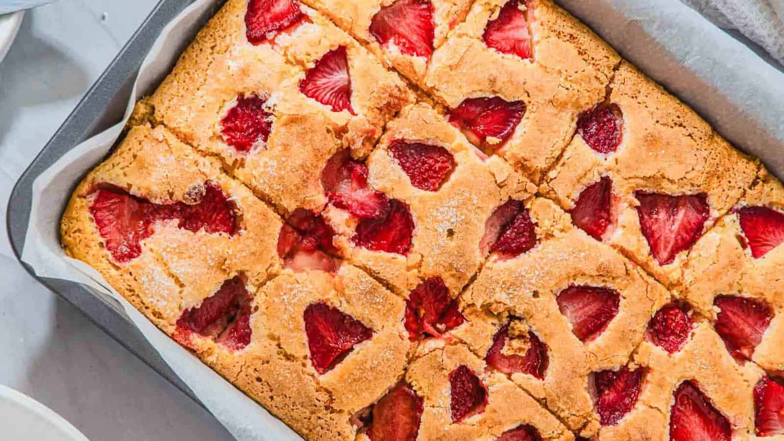 A slice of cake topped with strawberry sauce and a scoop of vanilla ice cream sits on a white plate, with a halved fresh strawberry beside it. A baking tray and whole strawberries are in the background.
