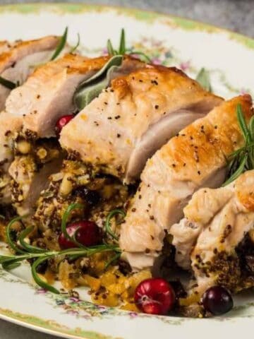 Sliced stuffed chicken breast served on a floral-patterned oval platter, garnished with fresh rosemary, sage leaves, and whole cranberries. The stuffing includes diced vegetables and herbs.