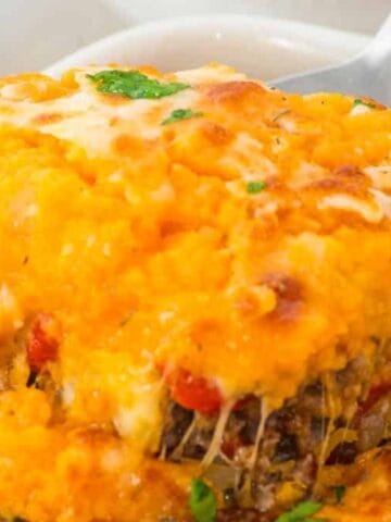 A close-up of a baked casserole with a golden, cheesy mashed potato topping and a visible vegetable and meat filling beneath, served in a white dish with a serving spoon.
