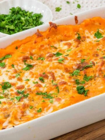 A baked sweet potato casserole topped with melted cheese and chopped parsley, served in a white rectangular dish on a wooden board. Small bowls of fresh herbs and seasonings are visible in the background.