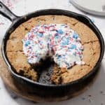 A large chocolate chip cookie cake baked in a cast iron skillet, topped with whipped cream and red, white, and blue star-shaped sprinkles—perfect for the 4th of July. A slice is missing, revealing the cookie’s soft interior.