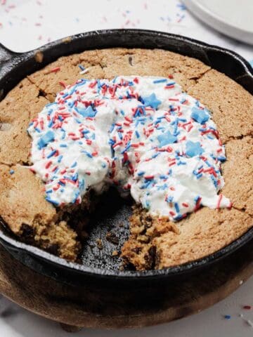 A large chocolate chip cookie cake baked in a cast iron skillet, topped with whipped cream and red, white, and blue star-shaped sprinkles—perfect for the 4th of July. A slice is missing, revealing the cookie’s soft interior.