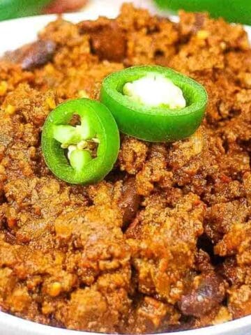 A white bowl filled with chili made of ground meat and beans, garnished with two slices of fresh green jalapeño pepper. The background includes part of a green pepper and a blue-and-white cloth.