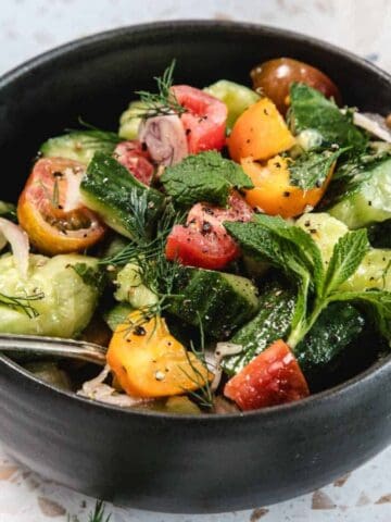 A black bowl filled with a colorful salad made of sliced cucumbers, red and yellow cherry tomatoes, fresh herbs, and a silver fork resting inside. The bowl is placed on a white speckled surface.
