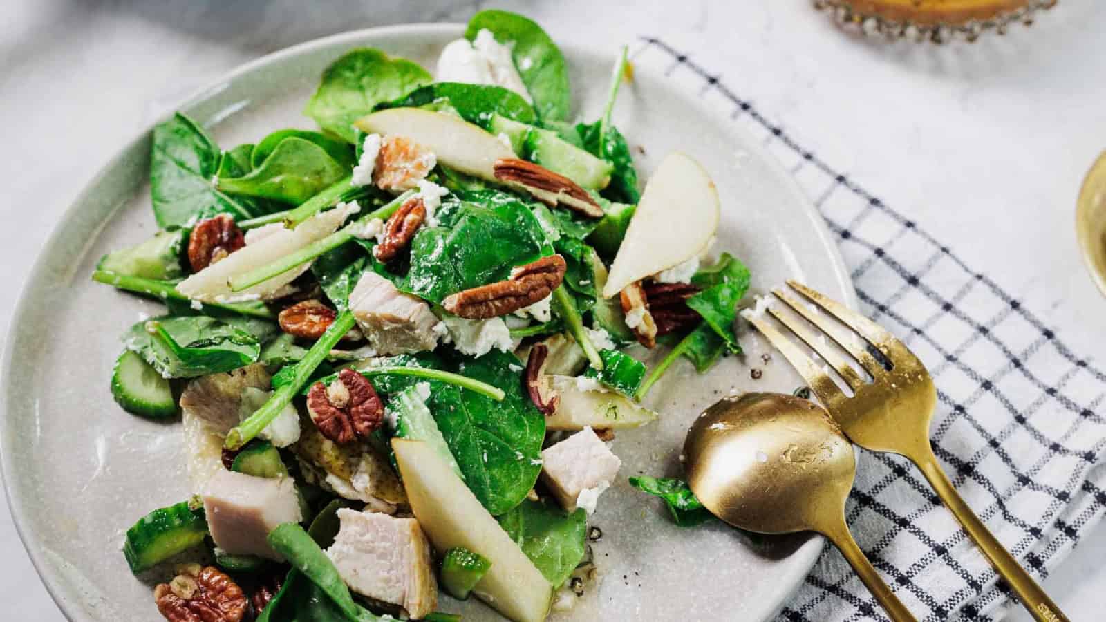 A plate of salad with spinach, slices of pear, pecans, chunks of chicken, cucumber, and cheese sits on a white surface next to a gold fork and spoon on a checkered napkin.