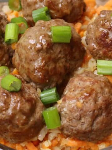 A clear bowl containing six cooked meatballs garnished with chopped green onions, served on a bed of diced carrots and cauliflower. The bowl is placed on a woven surface.