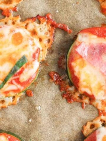 Sliced zucchini rounds topped with melted cheese, tomato sauce, and thin strips of basil on a sheet of parchment paper, with some cheese slightly browned and spreading around the slices.