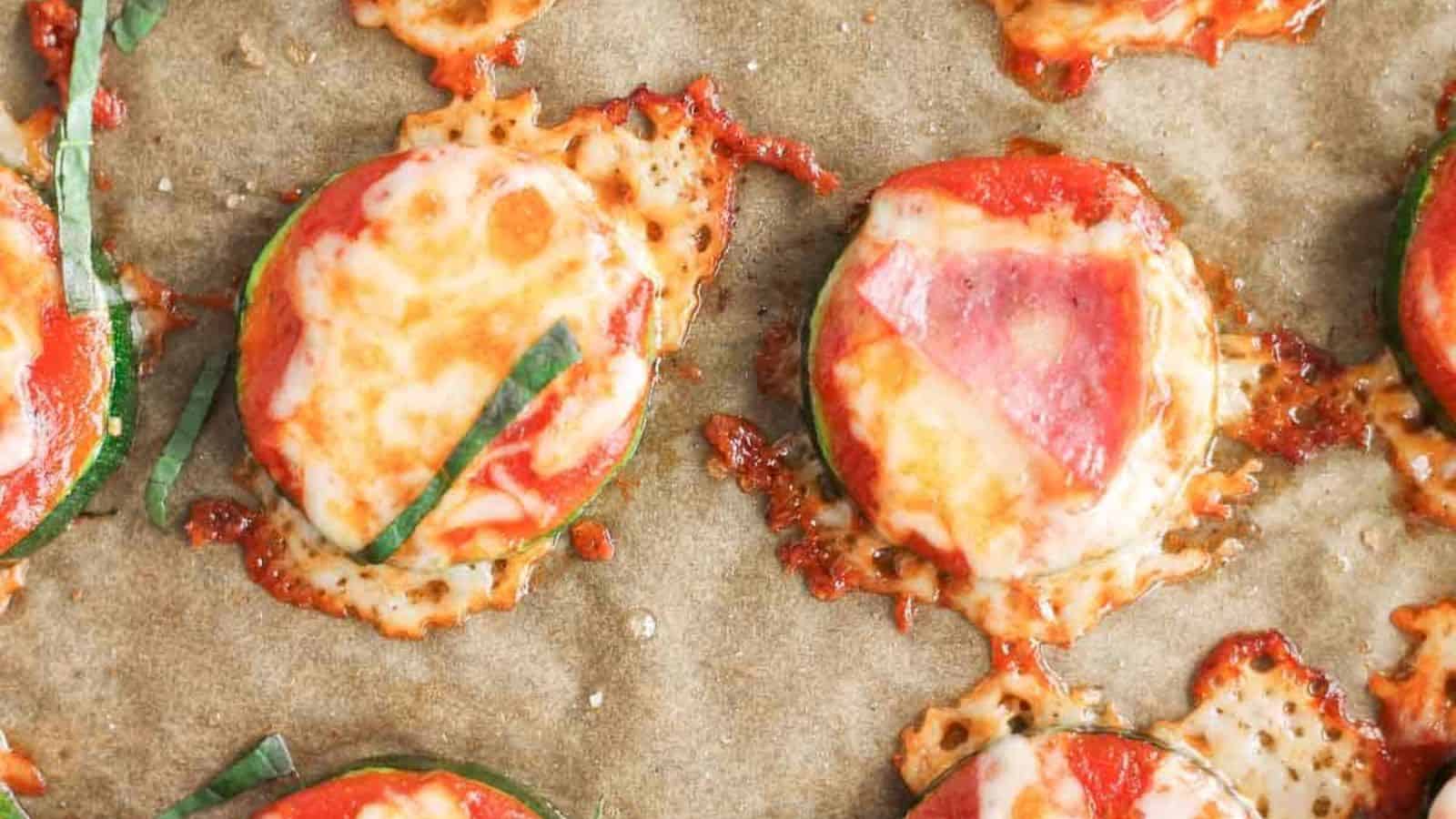Sliced zucchini rounds topped with melted cheese, tomato sauce, and thin strips of basil on a sheet of parchment paper, with some cheese slightly browned and spreading around the slices.