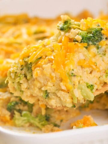 A wooden spoon scoops a serving of broccoli and cheese casserole from a white baking dish. The casserole is topped with melted cheddar cheese and contains visible pieces of broccoli and rice.
