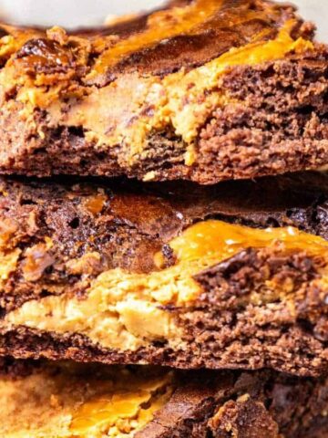 Three thick brownies with a marbled swirl of chocolate and peanut butter are stacked on top of each other. A spoon with peanut butter is visible in the lower right corner.