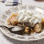 A serving of Peach Dump Cake made with yellow cake mix, topped with whipped cream, sits on a decorative plate with a spoon.