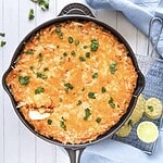 Cheesy Chicken Enchilada Dip baked in a skillet, topped with fresh herbs, served alongside tortilla chips and lime slices on a white surface.