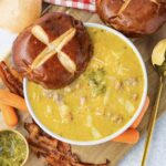 A bowl of instant pot cheeseburger soup, loaded with vegetables and ground meat, served with a pretzel roll, bacon, and carrots.