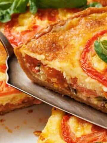 A slice of tomato and cheese quiche is being lifted from the whole quiche with a serving spatula.