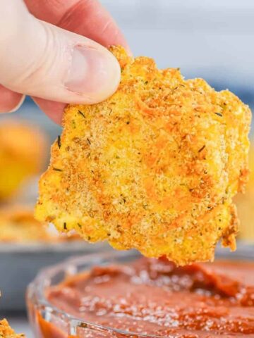 A hand dips a crispy, breaded slice into a bowl of red marinara sauce.