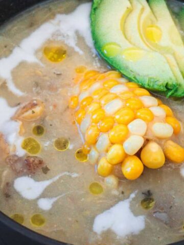 A bowl of ajiaco colombiano soup with chicken, corn on the cob, and avocado.