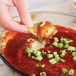 A hand dips a chip into a bowl of cheesy dip topped with salsa and chopped green onions.